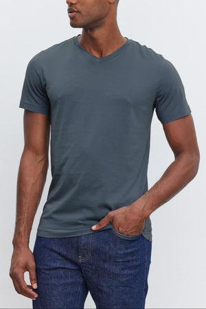 A man, showcasing everyday wear, is dressed in a fitted dark gray V-neck SAMSEN TEE by Velvet by Graham & Spencer and blue jeans. He stands against a plain light background, looking to the side with one hand in his pocket.