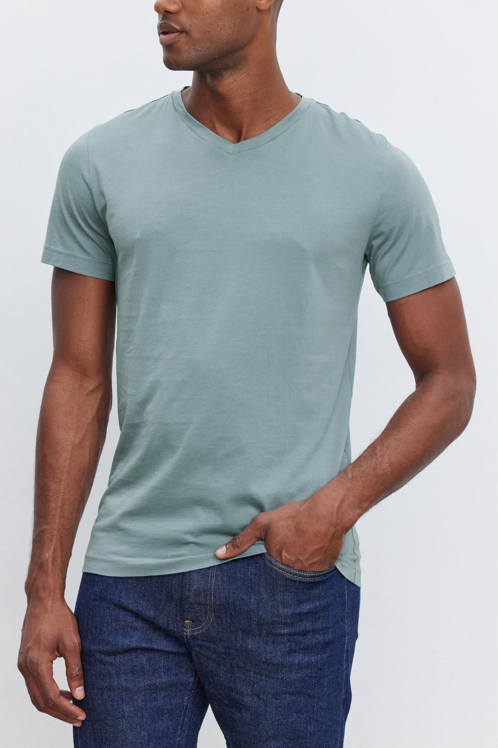 A man wearing a light green SAMSEN TEE made of whisper cotton knit by Velvet by Graham & Spencer and dark blue jeans stands against a plain light-colored background, looking to the side. This ensemble showcases stylish everyday wear with its comfortable fit and modern v-neckline.