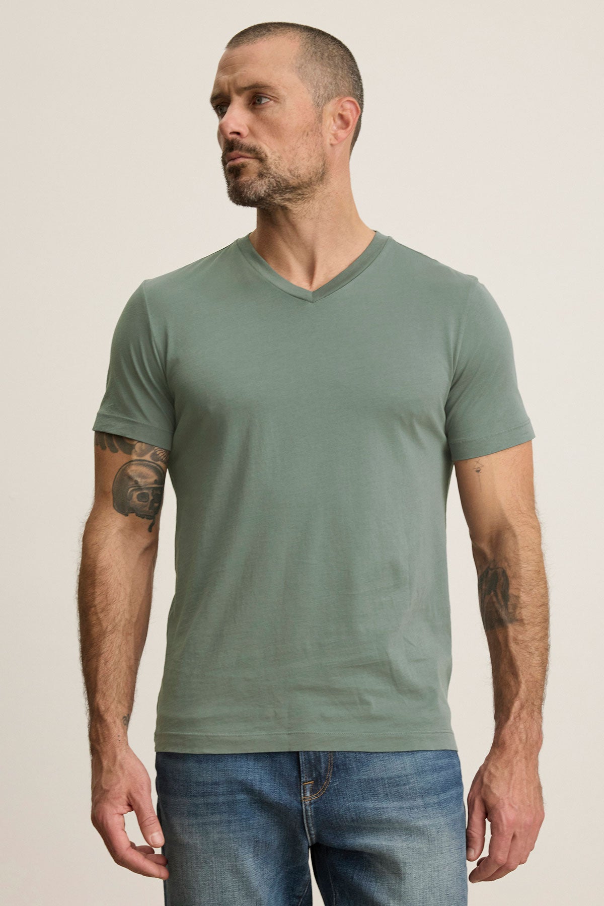 A man with short hair and tattoos on both arms stands against a neutral background, wearing the SAMSEN TEE by Velvet by Graham & Spencer in green V-neck Pima jersey with jeans.-38942945968321