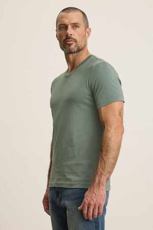 A bearded, short-haired man stands against a plain background wearing a light green Velvet by Graham & Spencer SAMSEN TEE with a v-neckline and blue jeans.