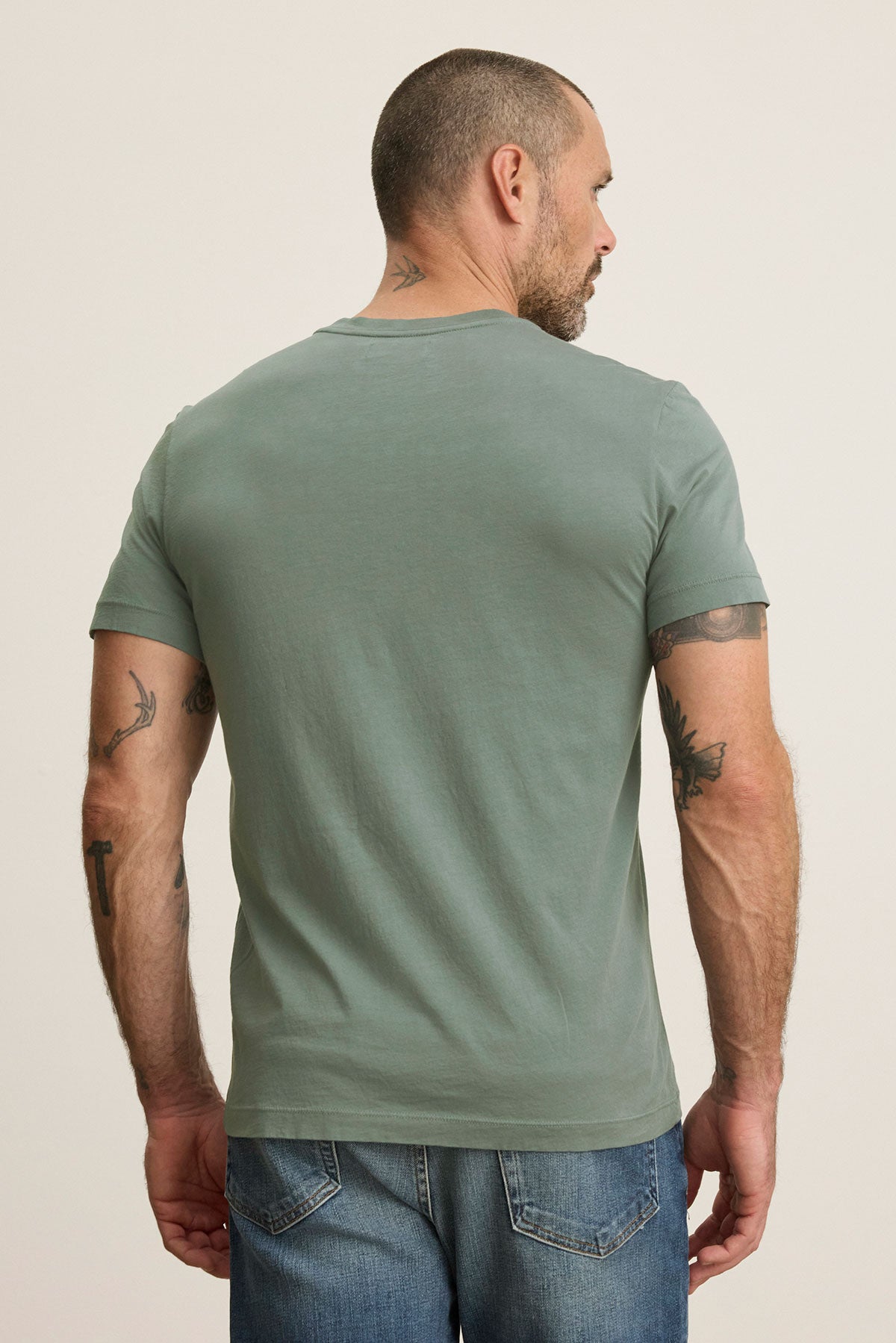 A person with short hair, viewed from behind, wears a light green Velvet by Graham & Spencer SAMSEN TEE with a v-neckline and blue jeans. Tattoos cover their arms and neck.