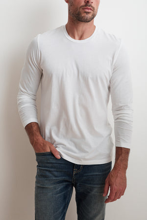A man in a plain white SKEETER TEE by Velvet by Graham & Spencer and blue jeans leans against a white wall, effortlessly embodying versatility with one hand casually in his pocket.