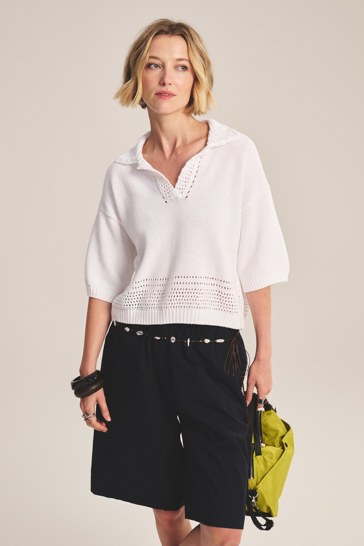 A woman wears the WINSLOW SWEATER POLO by Velvet by Graham & Spencer, a white pima cotton sweater with pointelle details, paired with black knee-length shorts and holding a yellow bag against a plain background.-41338498678977