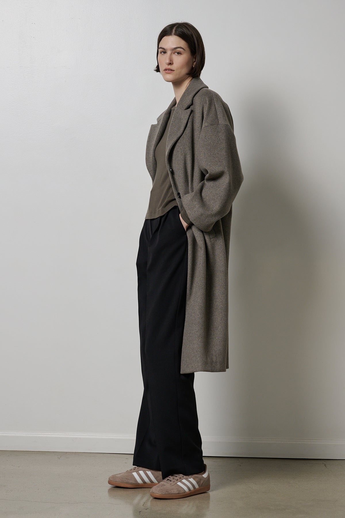 Person with short hair stands against a plain wall, wearing the HUNTINGTON COAT by Velvet by Jenny Graham, a wool blend gray coat with an oversized collar over a gray top, black pants, and brown sneakers with white stripes.-35766194143425