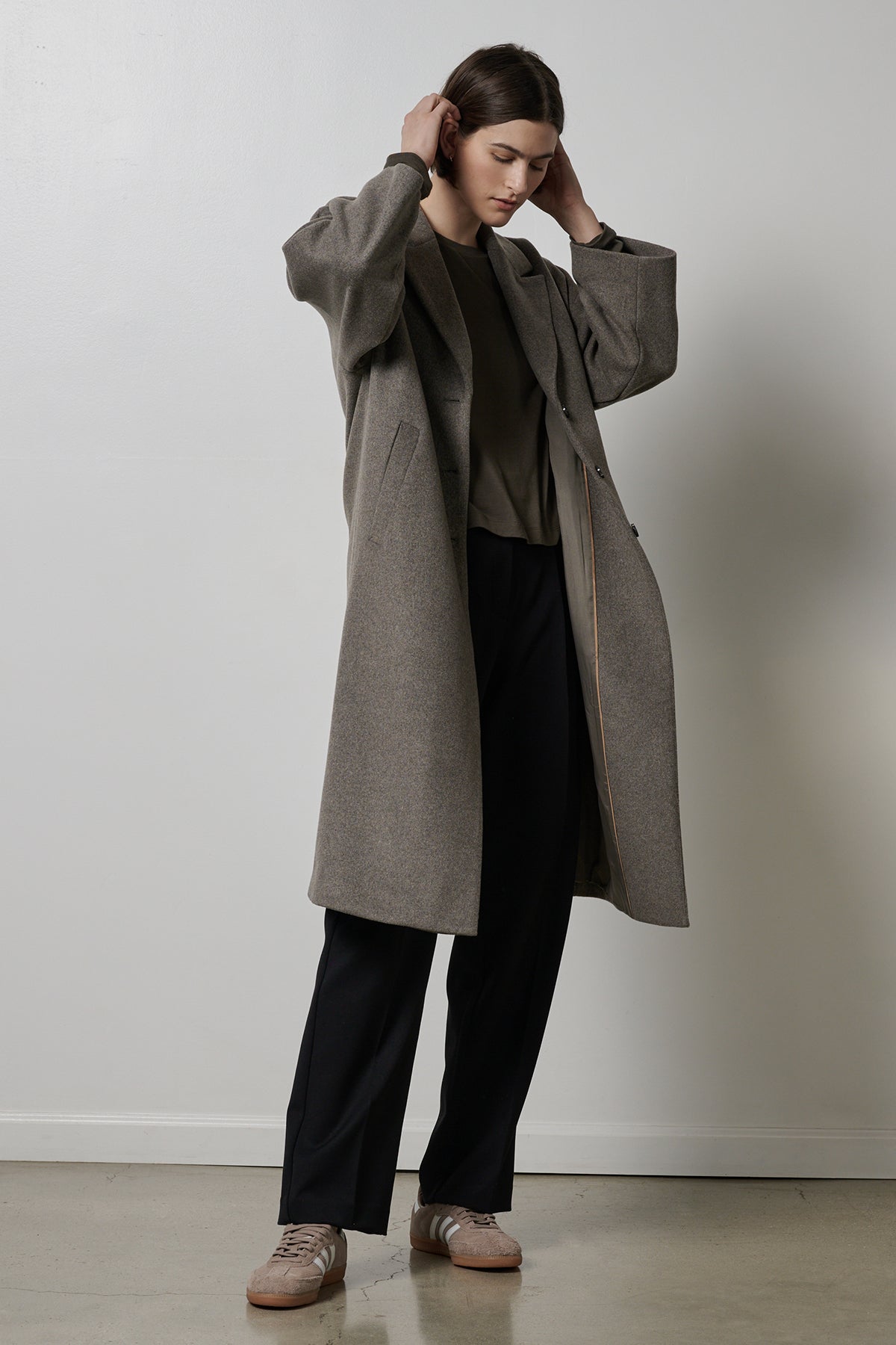 A person wearing a long grey, wool blend HUNTINGTON COAT by Velvet by Jenny Graham with an oversized collar over a dark shirt and black pants stands with hands adjusting their hair against a plain white wall background.-35766194176193