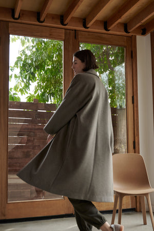 A person in a Velvet by Jenny Graham HUNTINGTON COAT with an oversized collar spins while standing indoors by large wooden-framed glass doors, with a tree visible outside.
