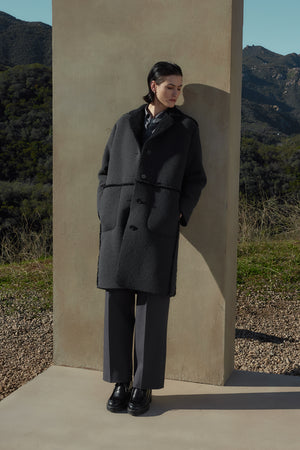 A person with short dark hair wearing the ROSEMEAD COAT by Velvet by Jenny Graham stands against a stone wall in an outdoor setting with mountains in the background.
