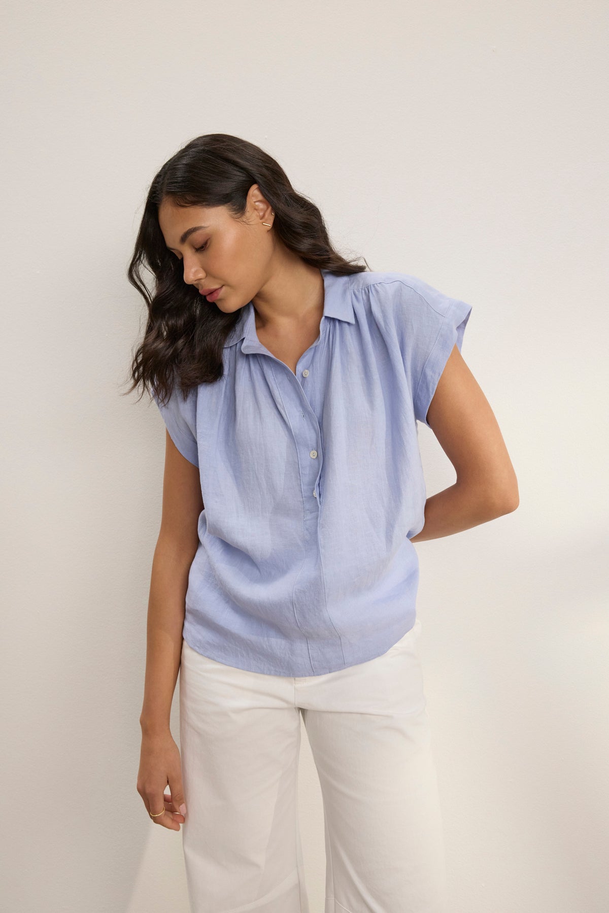 A woman wearing the ELOWEN LINEN TOP by Velvet by Graham & Spencer in light blue breathable linen and white pants stands against a plain background, looking down with one arm behind her back.-39123285016769