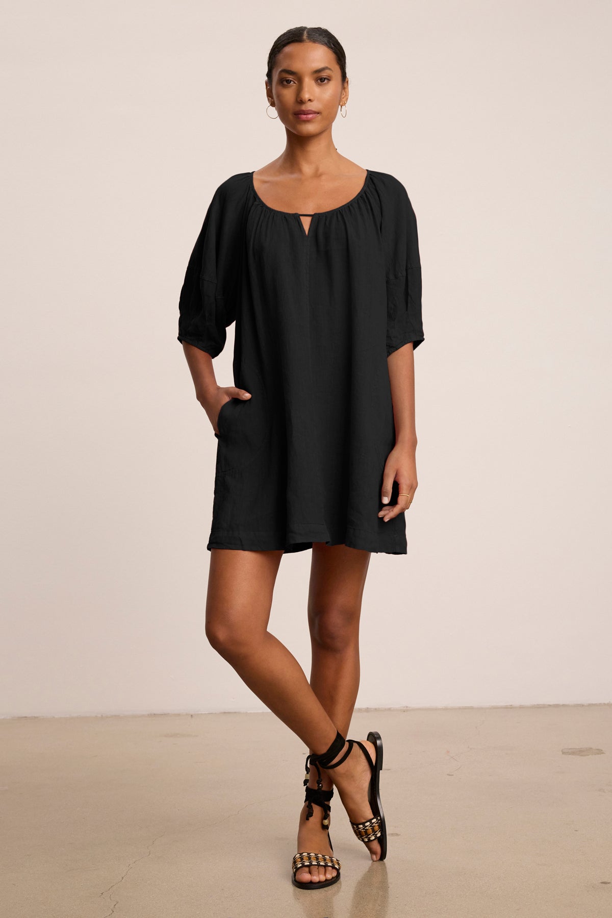 A woman stands indoors wearing the GRETA LINEN DRESS by Velvet by Graham & Spencer—a loose-fitting, black woven linen scoop neck dress with elbow-length sleeves. She has one hand in her pocket and looks at the camera.-39149302513857