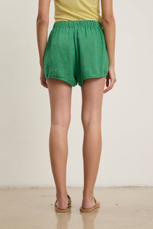 Person standing indoors, shown from the waist down, wearing a yellow top and the relaxed-fit JADEN LINEN SHORT by Velvet by Graham & Spencer in green—ideal for warm weather—paired with sandals.