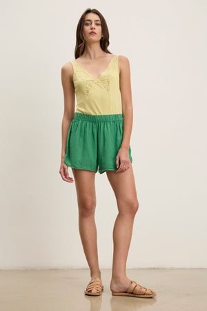 A woman stands indoors wearing a yellow sleeveless lace-trim top, tan sandals, and the JADEN LINEN SHORT by Velvet by Graham & Spencer—breathable green linen shorts perfect for warm-weather dressing.