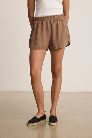 A person wearing a white tank top, black espadrille shoes, and Velvet by Graham & Spencer’s JADEN LINEN SHORT in brown with an elastic waist stands on a smooth floor against a plain background.