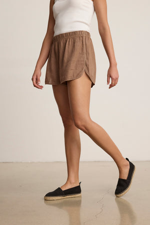 A person wears a white tank top, Velvet by Graham & Spencer's JADEN LINEN SHORT in brown with an elastic waist, and black espadrilles while standing on a polished floor against a plain background.