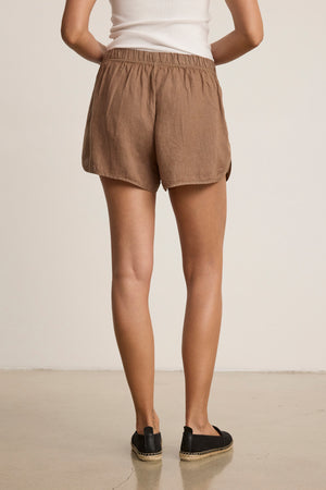 Person standing indoors facing away, wearing a white tank, black slip-ons, and the JADEN LINEN SHORT by Velvet by Graham & Spencer—relaxed-fit brown drawstring shorts in breathable linen, perfect for warm-weather style.