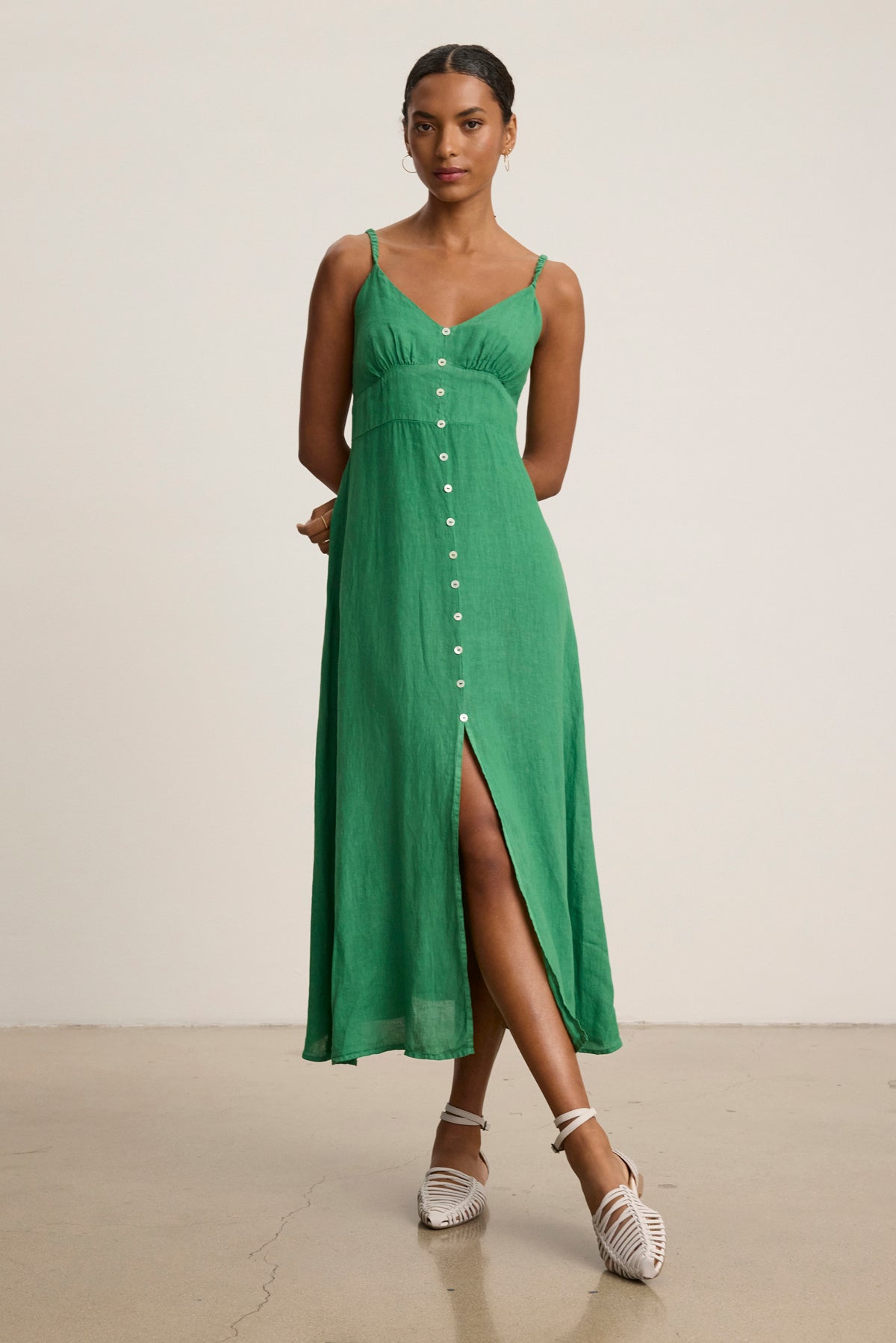 A woman wears the Velvet by Graham & Spencer JANET LINEN DRESS, a sleeveless green V-neck tank dress with front buttons, made of breathable linen, paired with white strappy shoes against a plain background.