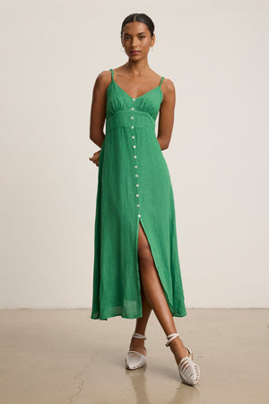 A woman wears the Velvet by Graham & Spencer JANET LINEN DRESS, a sleeveless green V-neck tank dress with front buttons, made of breathable linen, paired with white strappy shoes against a plain background.