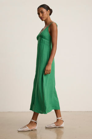 A woman stands sideways against a plain background wearing the Velvet by Graham & Spencer JANET LINEN DRESS, a sleeveless, ankle-length green breathable woven linen tank dress, paired with white woven sandals.
