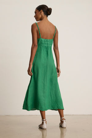 A woman stands indoors facing away, wearing the JANET LINEN DRESS by Velvet by Graham & Spencer—a sleeveless green woven linen tank dress—paired with white flat sandals.