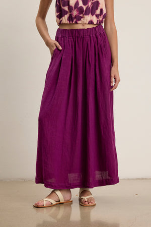 A person wearing the Velvet by Graham & Spencer LINDA LINEN SKIRT with an elastic waistband, a patterned sleeveless top, and beige sandals stands against a plain wall with one hand in their pocket.