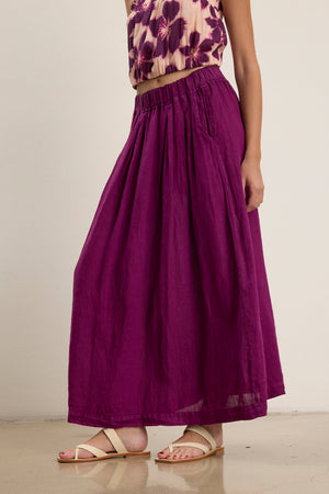 A person wears the LINDA LINEN SKIRT by Velvet by Graham & Spencer—a purple A-line maxi skirt—paired with a floral sleeveless top and beige sandals, standing indoors against a plain wall.