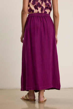 A person models the Velvet by Graham & Spencer LINDA LINEN SKIRT, a purple woven linen maxi with an A-line silhouette, paired with a patterned top and beige sandals, standing against a plain backdrop.