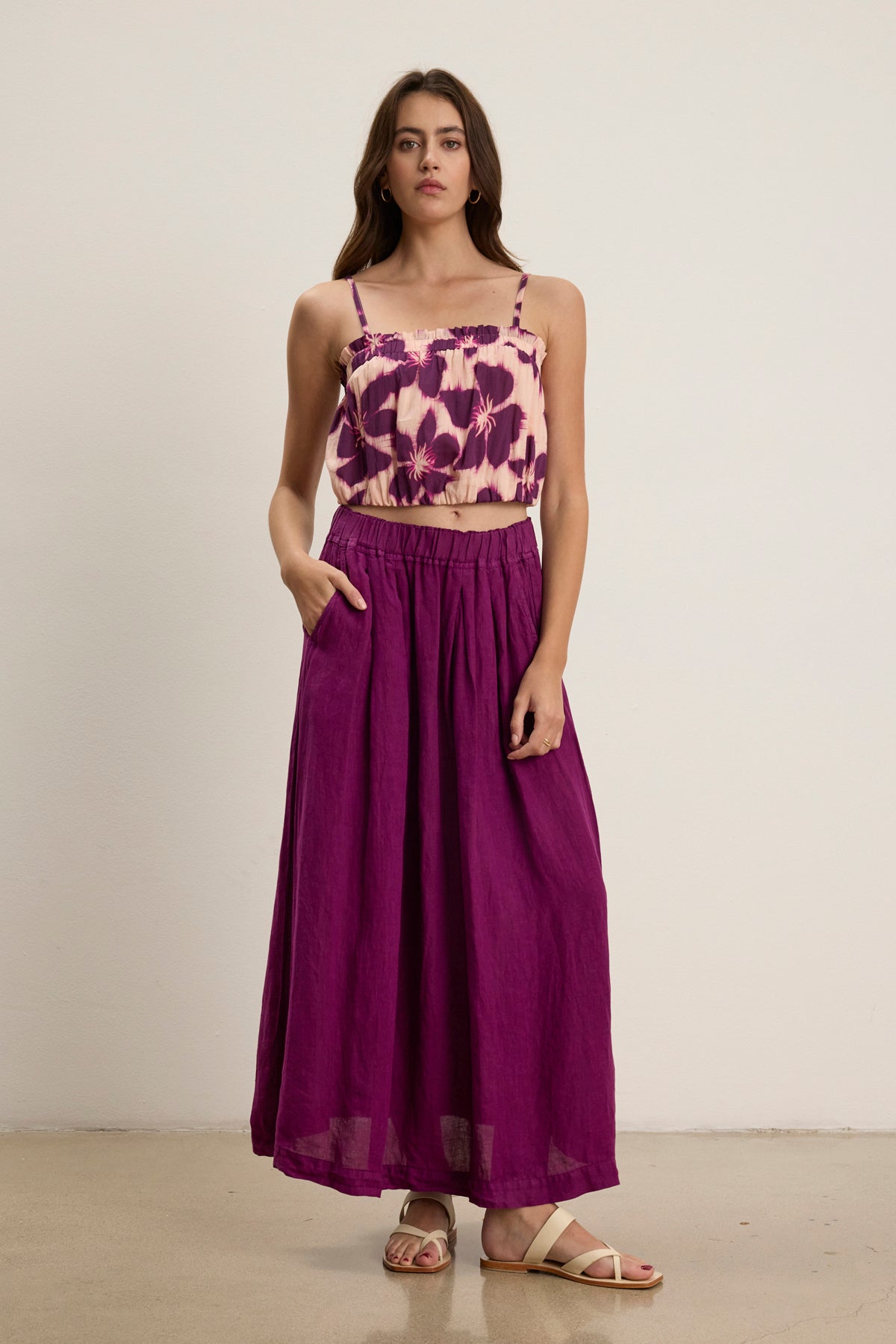 A woman stands against a plain background wearing a purple floral crop top, the LINDA LINEN SKIRT by Velvet by Graham & Spencer with an A-line silhouette and elastic waistband, and white sandals.-39111856160961