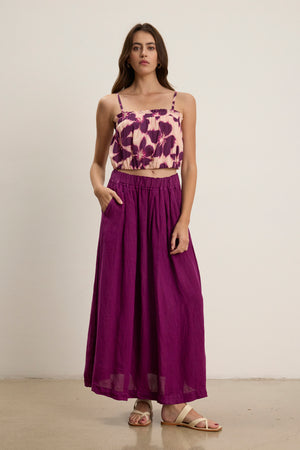 A woman stands against a plain background wearing a purple floral crop top, the LINDA LINEN SKIRT by Velvet by Graham & Spencer with an A-line silhouette and elastic waistband, and white sandals.