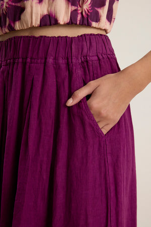 A person wearing the LINDA LINEN SKIRT by Velvet by Graham & Spencer in purple, featuring woven linen fabric, an elastic waistband, and a hand in the side pocket.