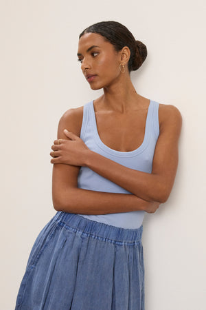 A woman wearing the Velvet by Graham & Spencer ZOEY TANK TOP in light blue stands with arms crossed against a plain wall, pairing the ribbed cotton scoop-neck staple with a blue skirt for a versatile, timeless look.