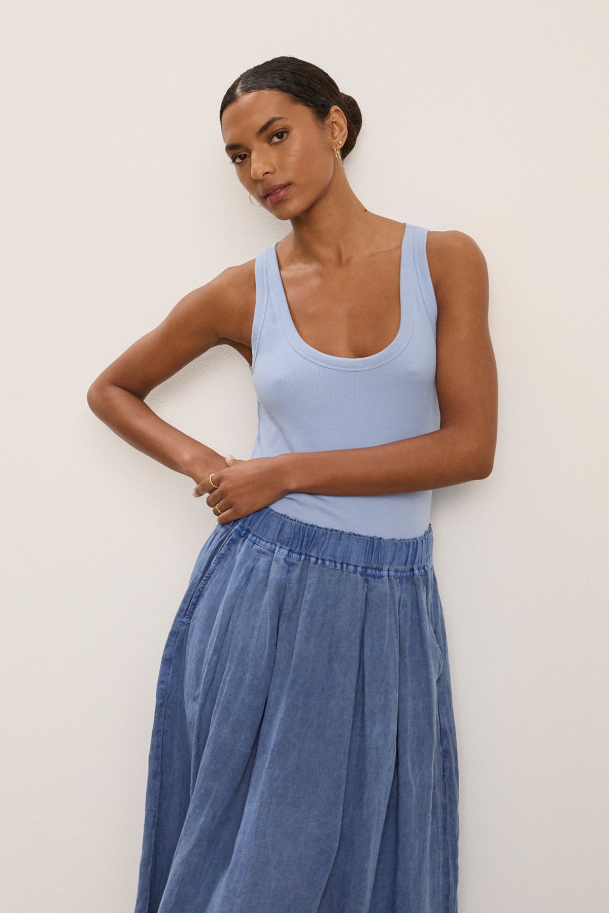 A woman poses against a plain light wall, wearing the ZOEY TANK TOP by Velvet by Graham & Spencer—a light blue ribbed cotton tank with a scoop neckline—paired with a flowy blue skirt and one arm on her waist.