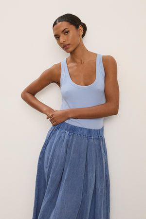 A woman poses against a plain light wall, wearing the ZOEY TANK TOP by Velvet by Graham & Spencer—a light blue ribbed cotton tank with a scoop neckline—paired with a flowy blue skirt and one arm on her waist.