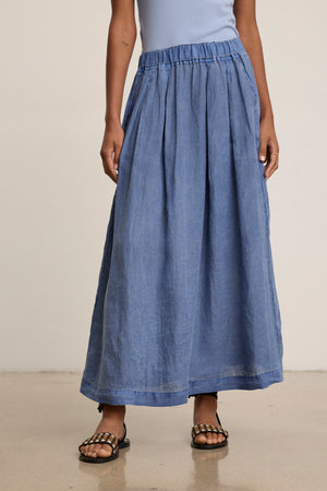 Person wearing a light blue sleeveless top, black sandals, and the LINDA LINEN SKIRT by Velvet by Graham & Spencer—a high-waisted blue A-line pleated maxi skirt—standing indoors on a polished floor.
