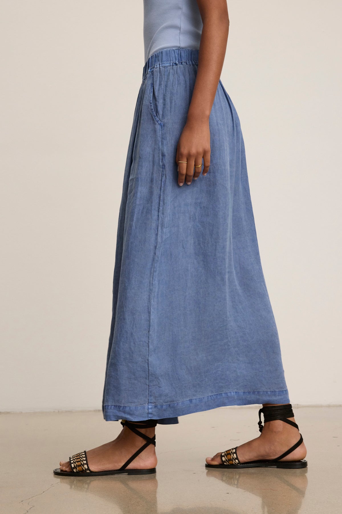 A person wearing a light blue top, Velvet by Graham & Spencer's LINDA LINEN SKIRT—a blue breathable A-line maxi skirt with pockets—and black strappy sandals is walking on a polished floor.