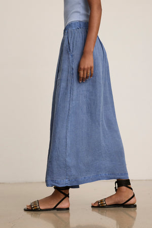 A person wearing a light blue top, Velvet by Graham & Spencer's LINDA LINEN SKIRT—a blue breathable A-line maxi skirt with pockets—and black strappy sandals is walking on a polished floor.