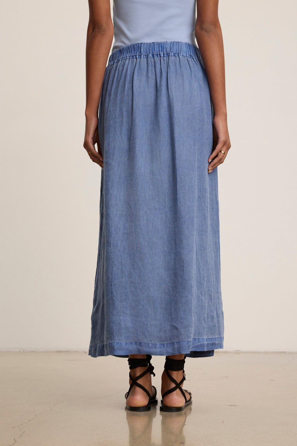 A person in a light blue sleeveless top, the LINDA LINEN SKIRT by Velvet by Graham & Spencer, and black sandals stands with their back to the camera against a neutral background.