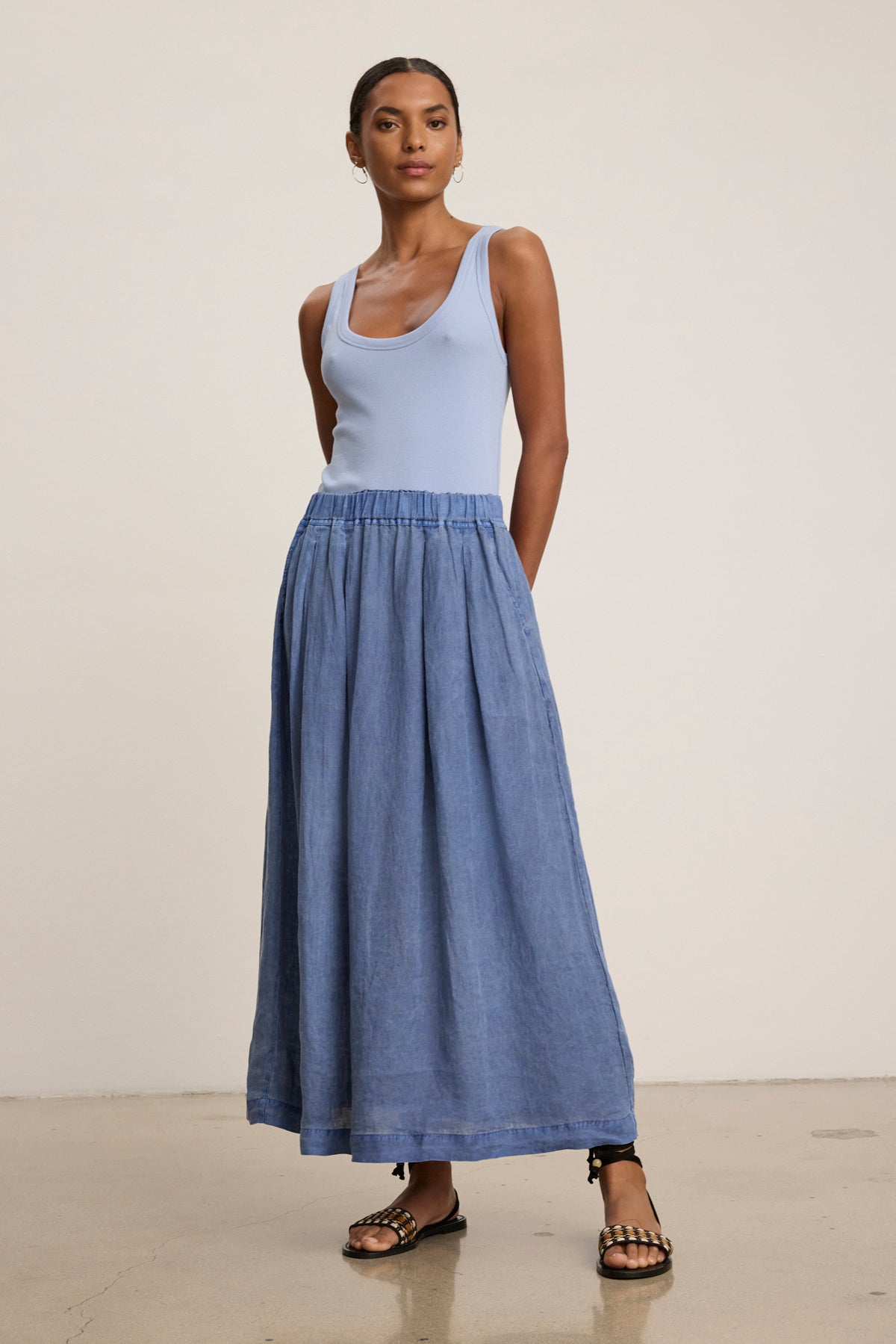 A woman stands against a neutral background wearing a light blue tank top, the LINDA LINEN SKIRT by Velvet by Graham & Spencer in blue with an A-line silhouette, and black sandals with gold accents.