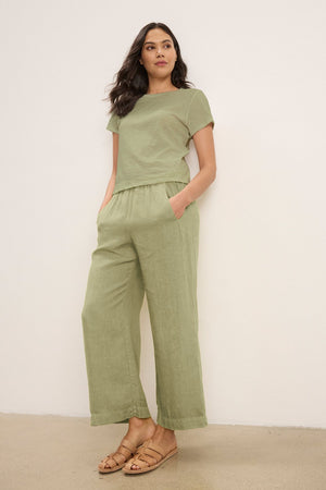 A woman stands against a plain wall wearing Velvet by Graham & Spencer's LOLA LINEN PANT in light green, paired with a matching short-sleeve top and tan sandals, her hands tucked in the pants' pockets.