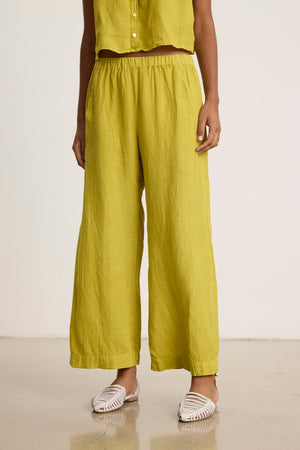 A person wearing the LOLA LINEN PANT by Velvet by Graham & Spencer, featuring an elastic waist in yellow, with a matching sleeveless top and white woven flats, stands on a polished floor against a plain background.