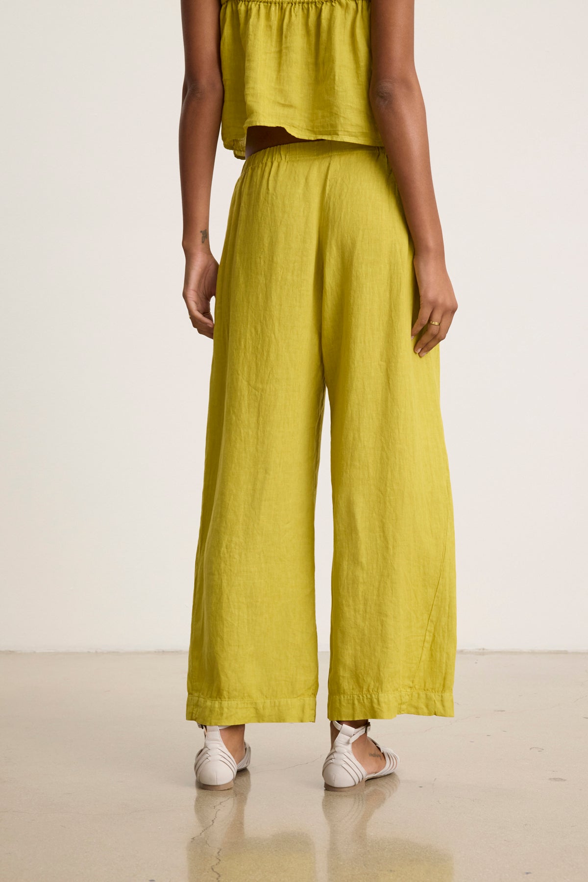 A person stands indoors facing away, wearing Velvet by Graham & Spencer's LOLA LINEN PANT in bright yellow-green with a matching top and white sandals.-39119919022273