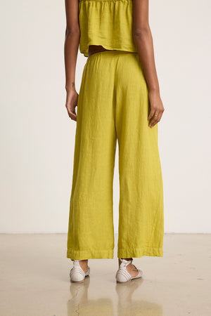 A person stands indoors facing away, wearing Velvet by Graham & Spencer's LOLA LINEN PANT in bright yellow-green with a matching top and white sandals.