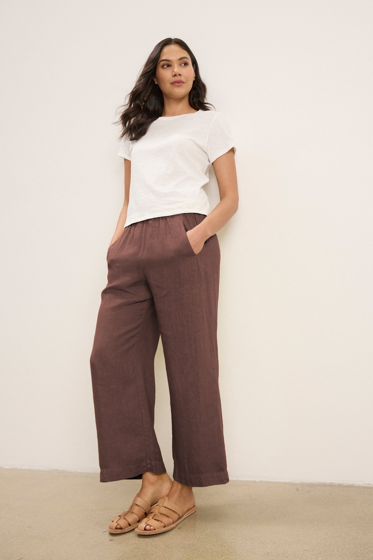 A woman stands against a light wall wearing Velvet by Graham & Spencer’s white tee and the LOLA LINEN PANT—loose brown pants with an elastic waist and relaxed leg—plus tan sandals, hands in pockets.-39333430919361