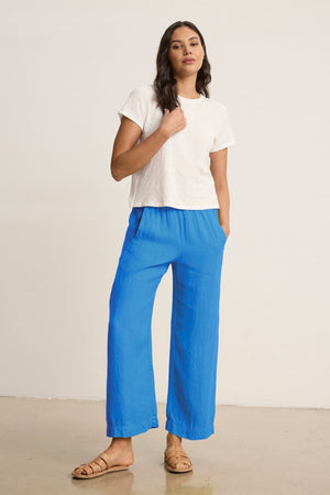 A woman stands indoors against a plain light background, wearing a white t-shirt, tan sandals, and Velvet by Graham & Spencer's LOLA LINEN PANT in bright blue with an elastic waist.