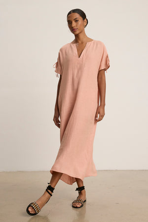 A woman wears the MAEVE LINEN DRESS by Velvet by Graham & Spencer, a loose-fitting light pink linen dress with a v-neckline and short sleeves, paired with black strappy sandals—perfect for casual outings.