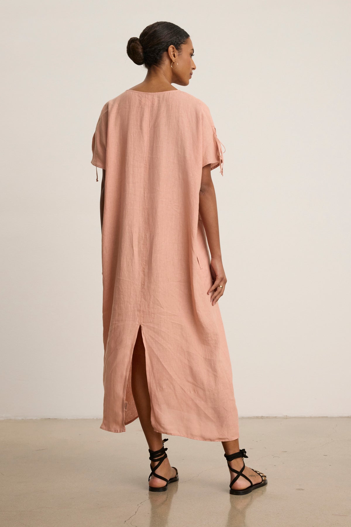 A woman indoors faces away, wearing the MAEVE LINEN DRESS by Velvet by Graham & Spencer—a loose, light pink linen caftan with a v-neckline and back slit—paired with black strappy sandals for casual outings.-39148737626305