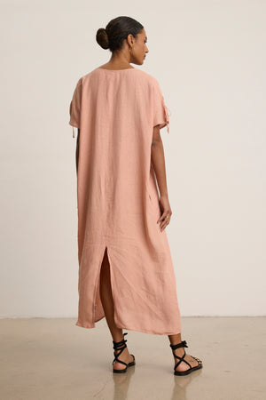 A woman indoors faces away, wearing the MAEVE LINEN DRESS by Velvet by Graham & Spencer—a loose, light pink linen caftan with a v-neckline and back slit—paired with black strappy sandals for casual outings.