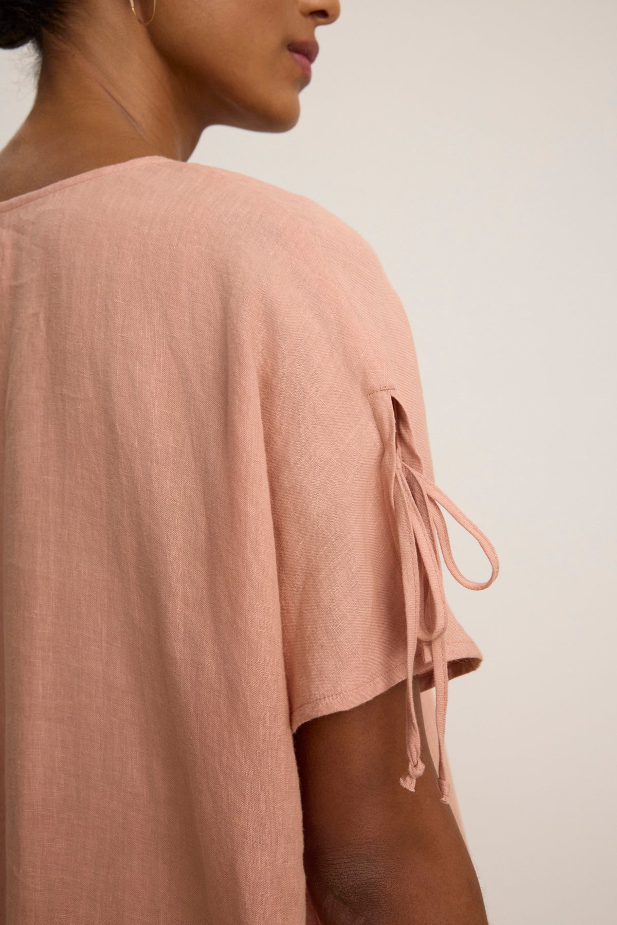 A close-up of a person wearing the MAEVE LINEN DRESS by Velvet by Graham & Spencer, featuring short sleeves with tied details and a v-neckline. The light pink shade and plain background highlight this stylish, casual look.-39148737593537