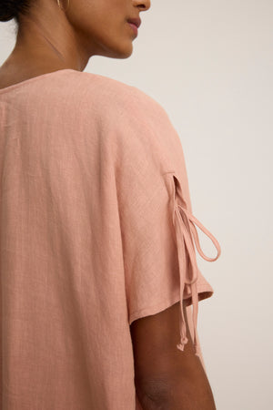 A close-up of a person wearing the MAEVE LINEN DRESS by Velvet by Graham & Spencer, featuring short sleeves with tied details and a v-neckline. The light pink shade and plain background highlight this stylish, casual look.