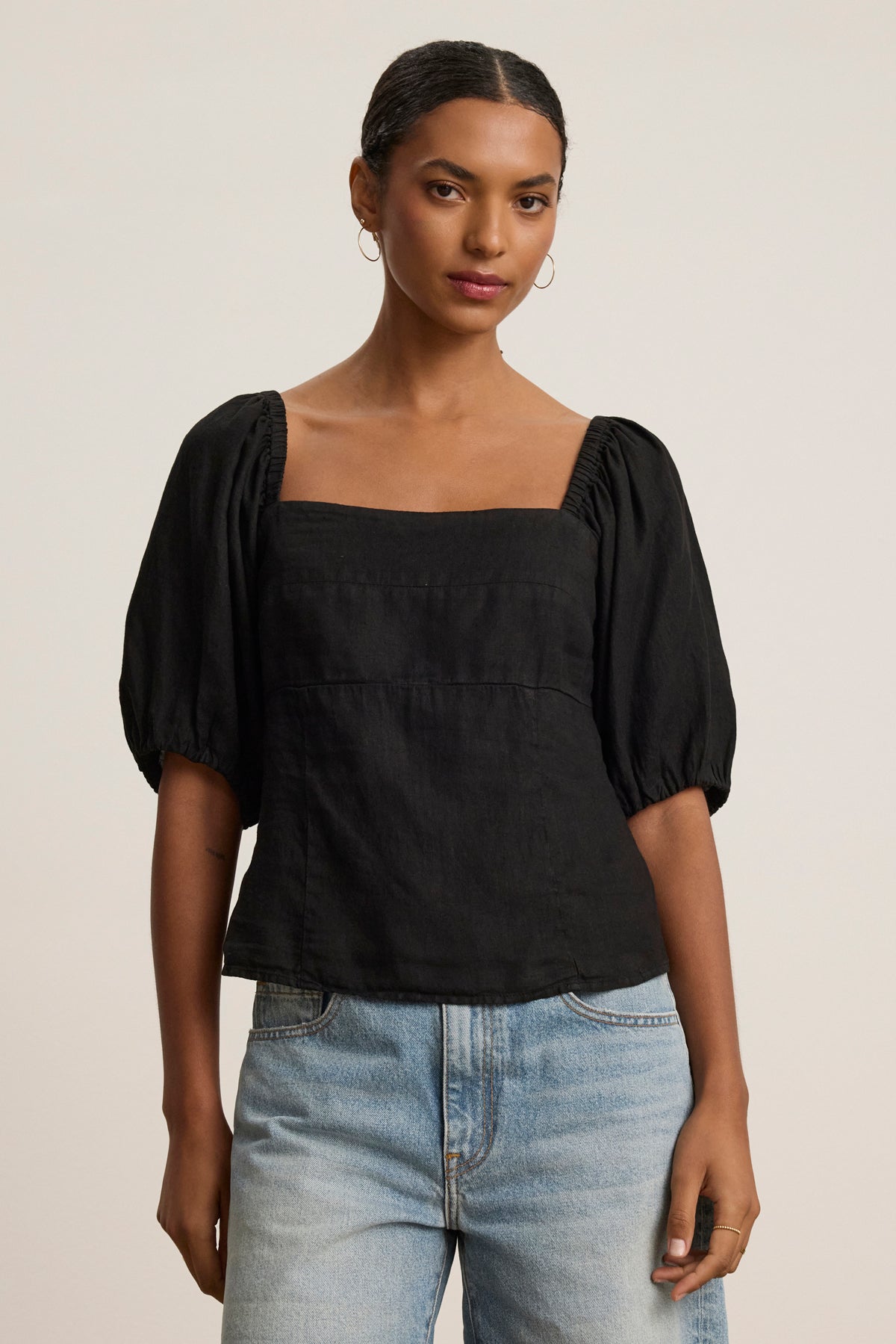 Someone stands against a plain background wearing the OLIVIA LINEN TOP by Velvet by Graham & Spencer—a black puff-sleeve top with a square neckline—paired with light blue jeans.