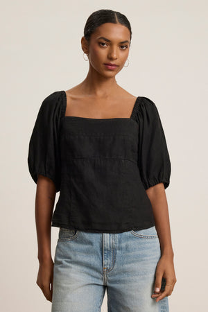 Someone stands against a plain background wearing the OLIVIA LINEN TOP by Velvet by Graham & Spencer—a black puff-sleeve top with a square neckline—paired with light blue jeans.