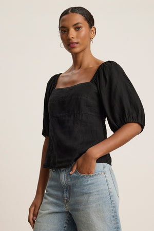 A woman in the OLIVIA LINEN TOP by Velvet by Graham & Spencer—a black, square-neck, puff sleeve cropped blouse—pairs it with light blue jeans, standing with one hand in her pocket against a plain background.